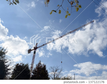 Crane above trees under blue sky. Industrial power, progress, and continuous city growth captured through construction symbolism. Crane above trees under blue sky. Industrial power, progress, and continuous city growth captured through construction symbolism. 137122176