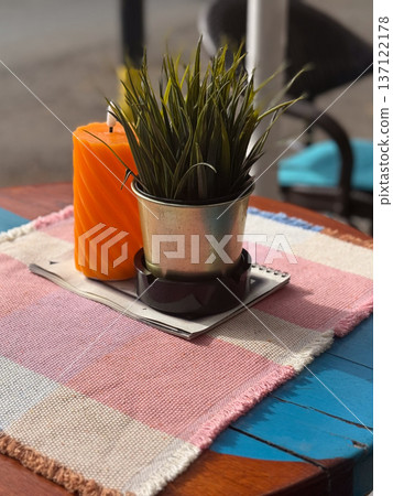 Outdoor cafe table with candle and potted plant on colorful fabric. Warmth, creativity and cozy leisure in street cafe ambiance. Outdoor cafe table with candle and potted plant on colorful fabric. Warmth, creativity and cozy leisure in street cafe ambiance. 137122178