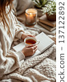 Young caucasian woman writing in a book and holding a mug of tea while relaxing wrapped in a cozy blanket indoors at home 137122892