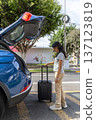 A teenager is standing next to a blue car with a suitcase in the trunk 137123819