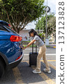 A teenager is standing next to a blue car and pulling a suitcase out of the trunk 137123828