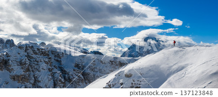 High resolution panoramic view of a ski mountaineer on a snowy ridge in the Dolomites with dramatic peaks and clouds, capturing winter adventure and vast alpine landscape. High resolution panoramic view of a ski mountaineer on a snowy ridge in the Dolomites with dramatic peaks and clouds, capturing winter adventure and vast alpine landscape. 137123848