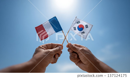 Hands holding small crossed french and south korean flags against a clear blue sky, celebrating the relationship between france and south korea Hands holding small crossed french and south korean flags against a clear blue sky, celebrating the relationship between france and south korea 137124175