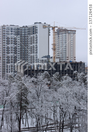 High Altitude Crane At Construction Site Of Modern Multi Storey Building Behind Small City Park Covered With Snow   137126458