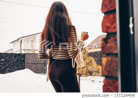 Back view of a young woman with a cup of coffee looking at a snowy backyard. Cozy winter morning atmosphere in a suburban house. Minimalist lifestyle photography of comfort and seasonal tranquility. 137126499