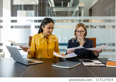 Diverse businesswomen reviewing paperwork and financial reports in the office, expressing confusion and frustration while dealing with complex data and potential problems Diverse businesswomen reviewing paperwork and financial reports in the office, expressing confusion and frustration while dealing with complex data and potential problems 137127034