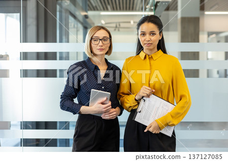 Two confident businesswomen standing in a contemporary office setting, holding a digital tablet and clipboard, representing collaboration, professionalism, and partnership in a corporate environment Two confident businesswomen standing in a contemporary office setting, holding a digital tablet and clipboard, representing collaboration, professionalism, and partnership in a corporate environment 137127085