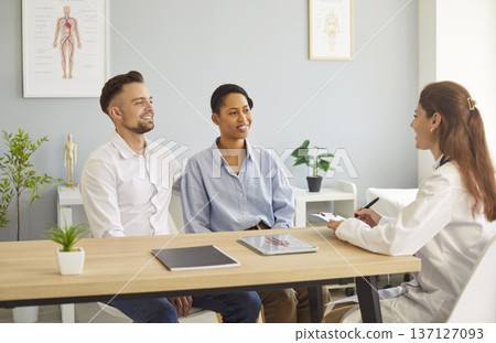 Smiling happy young couple sitting with family doctor therapist in medical clinic. Smiling happy young couple sitting with family doctor therapist in medical clinic. 137127093