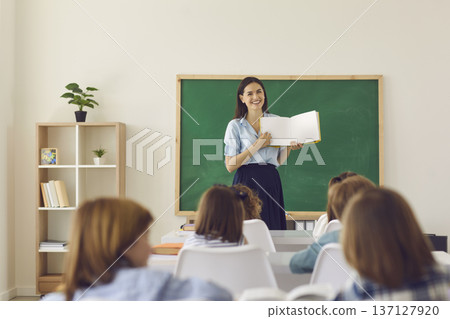 Smiling teacher explaining lesson with open book in classroom to engaged students Smiling teacher explaining lesson with open book in classroom to engaged students 137127920