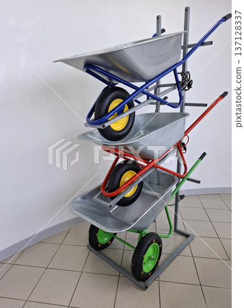 Metal wheelbarrows with blue, red, and green frames are arranged vertically in a hardware store. The display highlights different choices for those needing tools for yard work or projects. Metal wheelbarrows with blue, red, and green frames are arranged vertically in a hardware store. The display highlights different choices for those needing tools for yard work or projects. 137128337