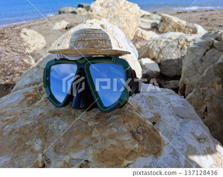 Diving goggles and a small straw hat rest on a sunlit rock by the ocean. Waves gently lap at the shore while the sky is bright and clear. This scene captures a moment of summer fun. Diving goggles and a small straw hat rest on a sunlit rock by the ocean. Waves gently lap at the shore while the sky is bright and clear. This scene captures a moment of summer fun. 137128436
