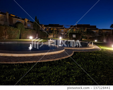 A curved swimming pool shines under warm lights in a quiet residential resort. The time is evening, and the surrounding area is still. 137128441