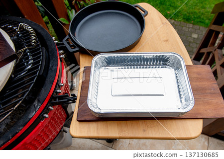 Ceramic grill with disposable aluminum trays and cast iron pan on side shelf outdoors 137130685