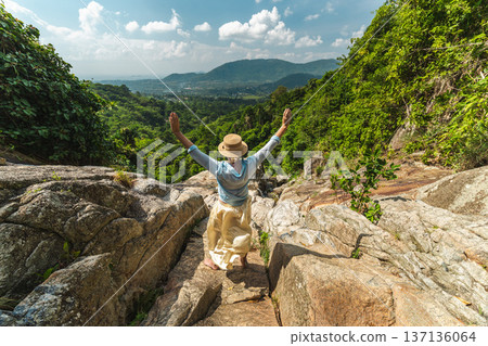 Young woman with raised arms standing on rocky path overlooking tropical valley and distant hills, summer travel freedom and adventure in lush green landscape Young woman with raised arms standing on rocky path overlooking tropical valley and distant hills, summer travel freedom and adventure in lush green landscape 137136064