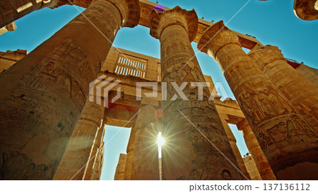 Ancient sandstone columns with carved hieroglyphs in Karnak Temple complex, Luxor Egypt, low angle view with dramatic sunlight and blue sky Ancient sandstone columns with carved hieroglyphs in Karnak Temple complex, Luxor Egypt, low angle view with dramatic sunlight and blue sky 137136112