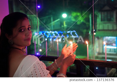 Beautiful Indian woman in white ethnic wear lighting candles on balcony during Diwali celebration Beautiful Indian woman in white ethnic wear lighting candles on balcony during Diwali celebration 137136376