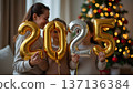 A family holds shiny golden balloons displaying 2025, standing in front of a decorated Christmas tree, radiating joy and excitement for the holidays and New Year celebrations 137136384