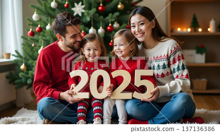 With a shining Christmas tree in the background, the family sits comfortably on the floor. They proudly hold cardboard 2025 numbers, their smiles radiating holiday joy 137136386