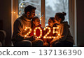 The family stands together in front of a large window framing a snowy yard. They hold glowing 2025 garlands, their faces lit with joy and the warmth of holiday spirit contrasting the frosty outdoors 137136390