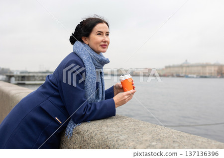 Woman sipping coffee leaning on riverside embankment over calm water in overcast weather. Woman sipping coffee leaning on riverside embankment over calm water in overcast weather. 137136396