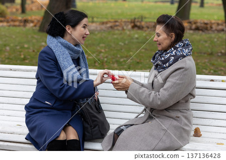 Mature women sitting on white wooden bench in autumn park engaged in friendly conversation wearing coats and scarves. Mature women sitting on white wooden bench in autumn park engaged in friendly conversation wearing coats and scarves. 137136428