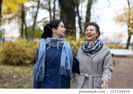 Mobility. Female friends strolling outdoors in cool season urban environment wearing coats and scarves communicating while moving through park area. Mobility. Female friends strolling outdoors in cool season urban environment wearing coats and scarves communicating while moving through park area. 137136430