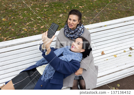 Two mature women sitting on white bench in autumn park taking selfie with smartphone smiling and posing together. Two mature women sitting on white bench in autumn park taking selfie with smartphone smiling and posing together. 137136432