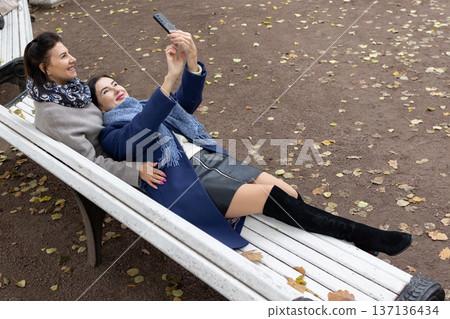 Female friends in coats and scarves embracing while photographing themselves outdoors during cool seasonal daytime. Female friends in coats and scarves embracing while photographing themselves outdoors during cool seasonal daytime. 137136434