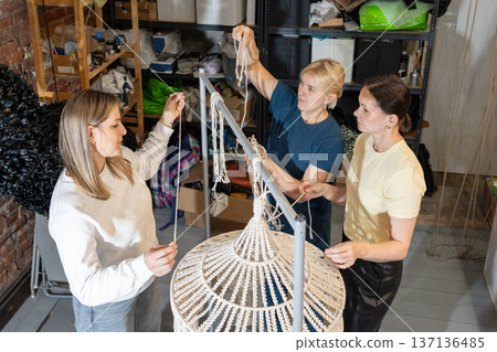Teaching mentor assisting women during hands on masterclass creating large woven pendant lamps from metal frames and rope. Teaching mentor assisting women during hands on masterclass creating large woven pendant lamps from metal frames and rope. 137136485