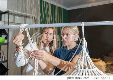 Guiding instructor demonstrating weaving technique while participants assemble decorative chandelier frames with cords in studio. Guiding instructor demonstrating weaving technique while participants assemble decorative chandelier frames with cords in studio. 137136487