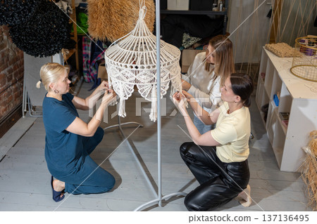 Guiding instructor demonstrating weaving technique while participants assemble decorative chandelier frames with cords in studio. Guiding instructor demonstrating weaving technique while participants assemble decorative chandelier frames with cords in studio. 137136495