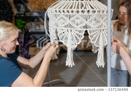 Teaching mentor assisting women during hands on masterclass creating large woven pendant lamps from metal frames and rope. Teaching mentor assisting women during hands on masterclass creating large woven pendant lamps from metal frames and rope. 137136499