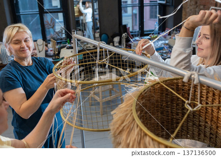 Teaching mentor assisting women during hands on masterclass creating large woven pendant lamps from metal frames and rope. Teaching mentor assisting women during hands on masterclass creating large woven pendant lamps from metal frames and rope. 137136500