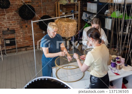 Teaching mentor assisting women during hands on masterclass creating large woven pendant lamps from metal frames and rope. Teaching mentor assisting women during hands on masterclass creating large woven pendant lamps from metal frames and rope. 137136505
