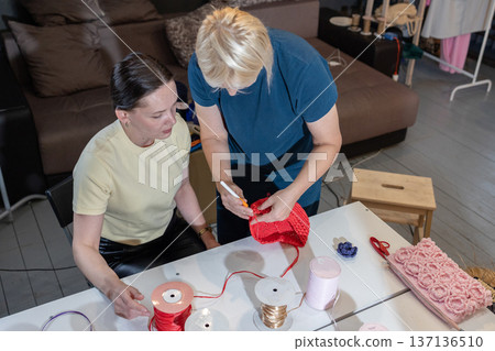 Three women crafting with colorful threads and crochet hooks at table in cozy workshop interior. Three women crafting with colorful threads and crochet hooks at table in cozy workshop interior. 137136510