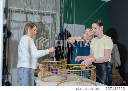 Teaching mentor assisting women during hands on masterclass creating large woven pendant lamps from metal frames and rope. Teaching mentor assisting women during hands on masterclass creating large woven pendant lamps from metal frames and rope. 137136513