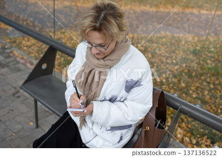 Mature woman with glasses holding notepad and pen outdoors near bench with autumn leaves on pavement. Mature woman with glasses holding notepad and pen outdoors near bench with autumn leaves on pavement. 137136522
