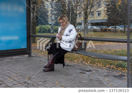 Mature woman with glasses holding notepad and pen outdoors near bench with autumn leaves on pavement. 137136523