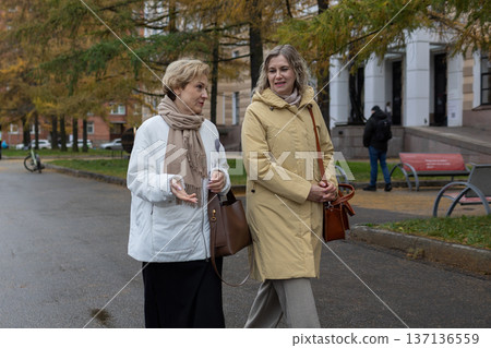 Mobility. Female friends strolling outdoors in cool season urban environment wearing coats and scarves communicating while moving through park area. Mobility. Female friends strolling outdoors in cool season urban environment wearing coats and scarves communicating while moving through park area. 137136559