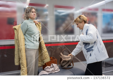 Two women taking off coats in cloakroom area of foyer placing outerwear in wardrobe during indoor arrival moment. Two women taking off coats in cloakroom area of foyer placing outerwear in wardrobe during indoor arrival moment. 137136572