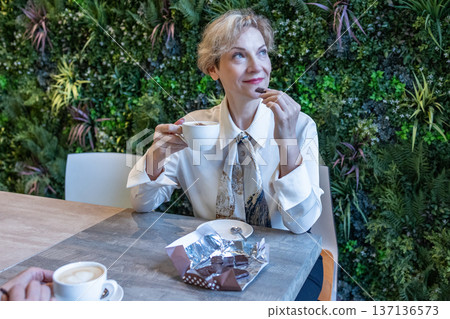 Contemplation. Woman sitting at cafe table holding cup of coffee with chocolate dessert while gazing thoughtfully aside in calm atmosphere. Contemplation. Woman sitting at cafe table holding cup of coffee with chocolate dessert while gazing thoughtfully aside in calm atmosphere. 137136573