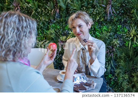 Two mature women sitting at wooden table in modern cafe holding cups of coffee and dessert engaged in friendly conversation. 137136574