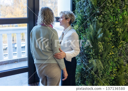 Two women appreciating contemporary art inside modern museum space sharing impressions and gestures while viewing framed paintings. Two women appreciating contemporary art inside modern museum space sharing impressions and gestures while viewing framed paintings. 137136580