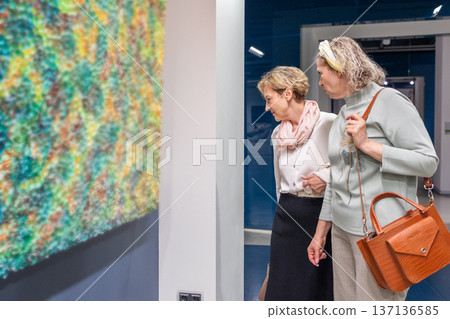 Women standing in gallery interior examining framed artwork and pointing during exhibition visit in quiet discussion. Women standing in gallery interior examining framed artwork and pointing during exhibition visit in quiet discussion. 137136585