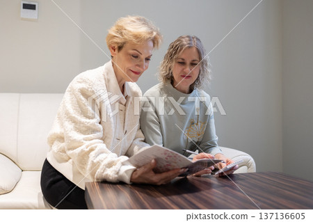 Two mature women sitting on light sofa examining open brochure on table discussing printed images in quiet interior space. Two mature women sitting on light sofa examining open brochure on table discussing printed images in quiet interior space. 137136605