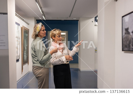 Women standing in gallery interior examining framed artwork and pointing during exhibition visit in quiet discussion. Women standing in gallery interior examining framed artwork and pointing during exhibition visit in quiet discussion. 137136615