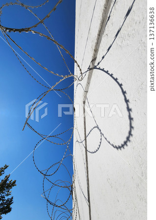 Barbed wire on white wall and blue sky with shadows from bright sun. High quality photo Barbed wire on white wall and blue sky with shadows from bright sun. High quality photo 137136638