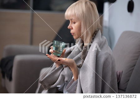 Medication intake. Blonde woman wrapped in blanket sitting on sofa holding pills in palm preparing to take medicine indoors during illness recovery. Medication intake. Blonde woman wrapped in blanket sitting on sofa holding pills in palm preparing to take medicine indoors during illness recovery. 137136872