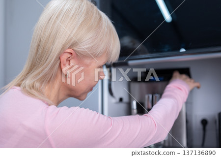Coffee preparation. Adult blonde woman operating espresso machine holding empty glass cup in bright kitchen interior with focus on routine 137136890