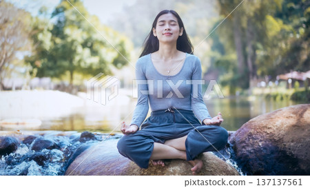 Young woman meditating cross-legged on rock by a calm river in sunny park, eyes closed, relaxed Young woman meditating cross-legged on rock by a calm river in sunny park, eyes closed, relaxed 137137561